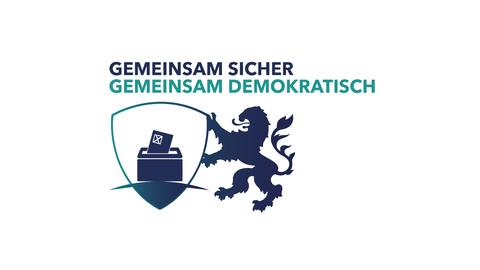 Ein aufsteigender Hessenlöwe der einen Schild mit abgebildeter Wahlbox präsentiert. Der Schriftzug lautet "Gemeinsam Sicher Gemeinsam Demokratisch"